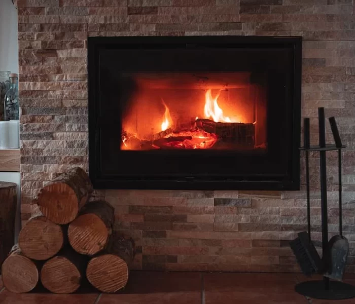 A cozy fireplace with burning logs is set into a stone wall. Firewood is stacked neatly beside it, along with fireplace tools and a small potted plant on a nearby shelf&mdash;perfect for relaxing after air duct services New Jersey.