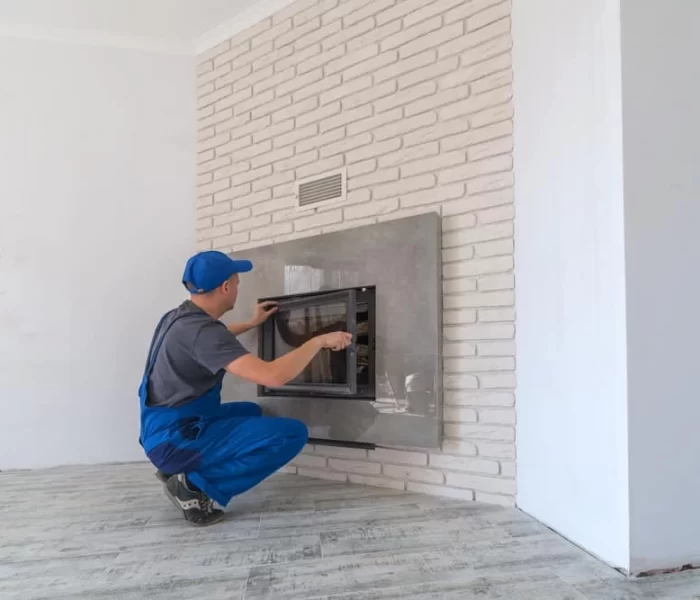 A worker in blue overalls and cap installs or repairs a glass fireplace door on a white brick wall in a modern, unfurnished room with light-colored wooden flooring, where air duct services New Jersey are also available.
