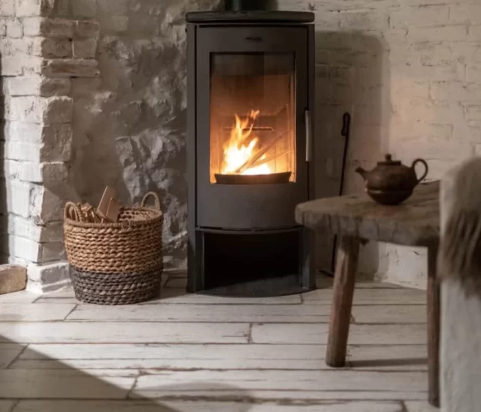 A cozy room with a lit wood-burning stove, a wicker basket of firewood, a rustic wooden table with a teapot, and whitewashed brick walls and floors&mdash;perfect after trusted air duct services New Jersey has to offer.