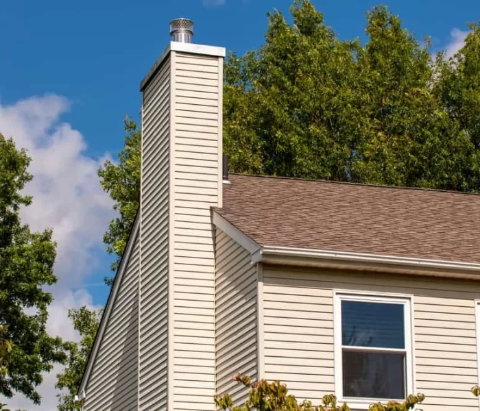 Beige house with a steep roof, vinyl siding, and a tall chimney against green trees and a partly cloudy blue sky. Two windows are visible&mdash;ideal for seeking air duct services New Jersey to keep your home fresh year-round.