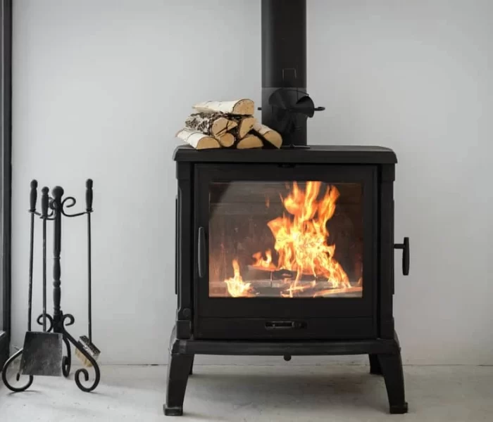A black wood-burning stove with a visible fire inside sits in a corner. Logs are stacked on top of the stove. Fireplace tools, including a poker and shovel, stand nearby&mdash;reminding you of the importance of air duct services New Jersey offers.